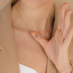 Someone wearing the Maia Emerald Necklace, a gold pendant featuring a small green emerald, gently holds it near their neck.