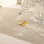 The Pave Heart Knot Ring, a gold promise ring with a knot design, rests on a light surface near a clear textured glass object—a romantic and meaningful gift.