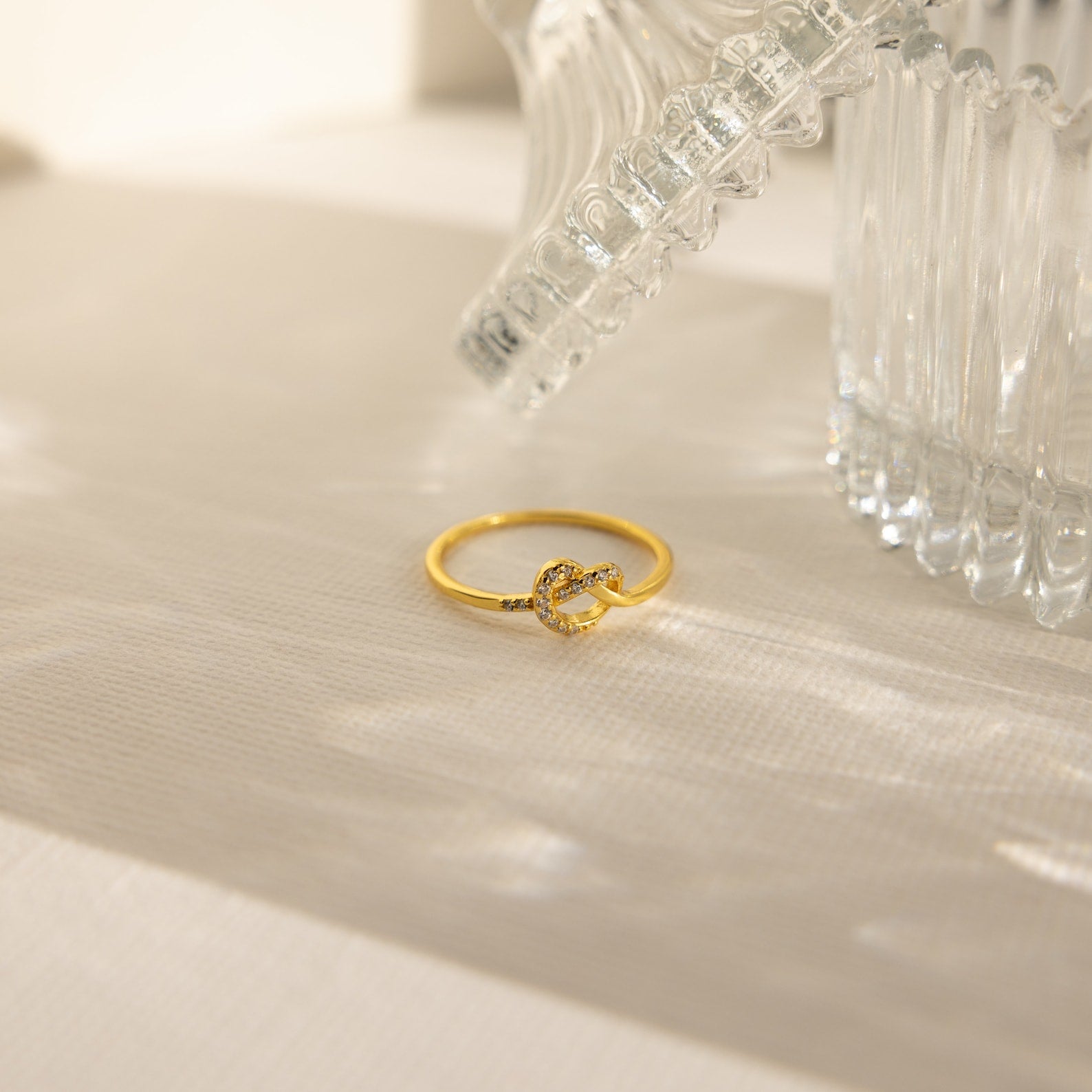 The Pave Heart Knot Ring, a gold promise ring with a knot design, rests on a light surface near a clear textured glass object—a romantic and meaningful gift.