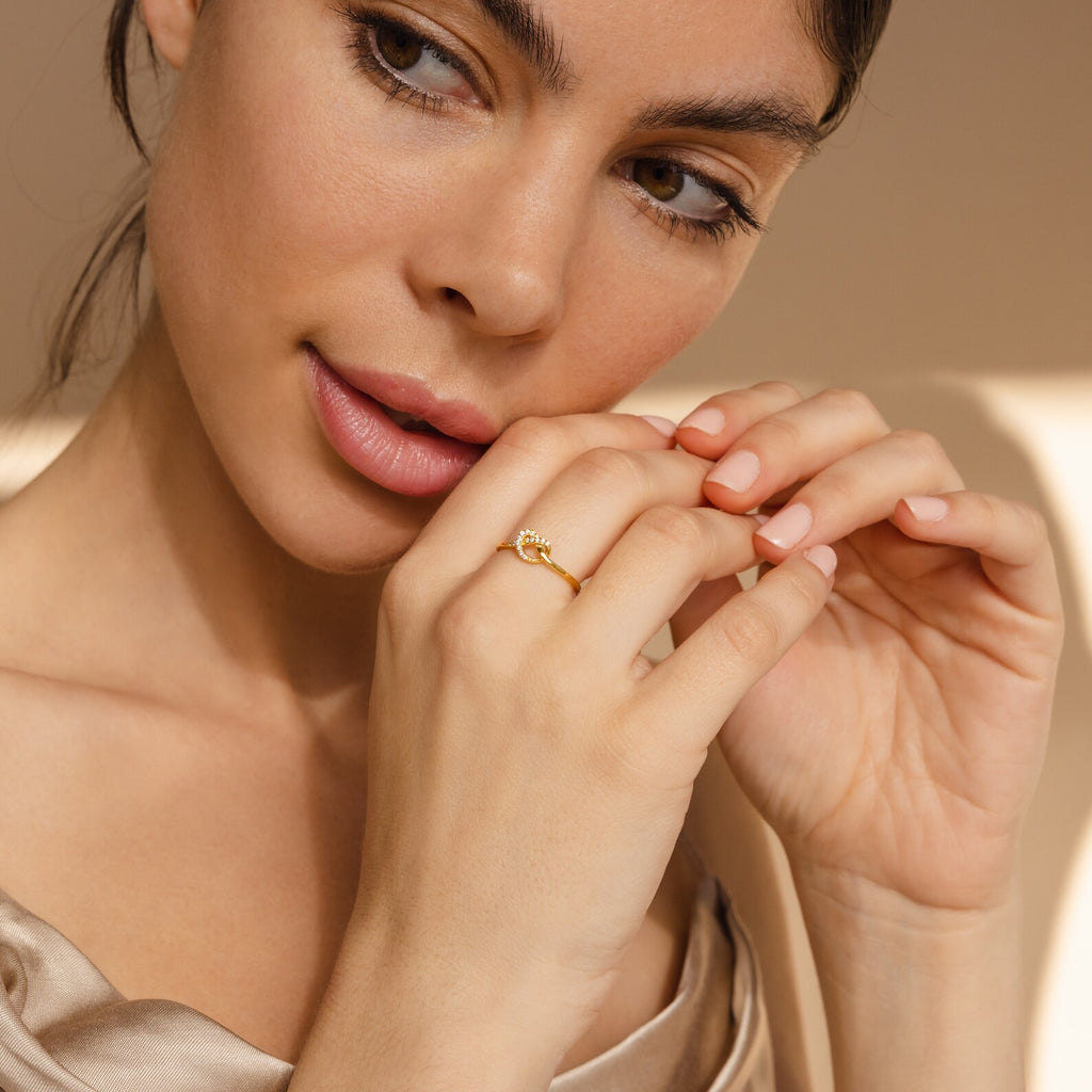 A woman with brown hair wears the Pave Heart Knot Ring, gently touching her face and hand, looking thoughtful—an elegant symbol of romance and a perfect gift.