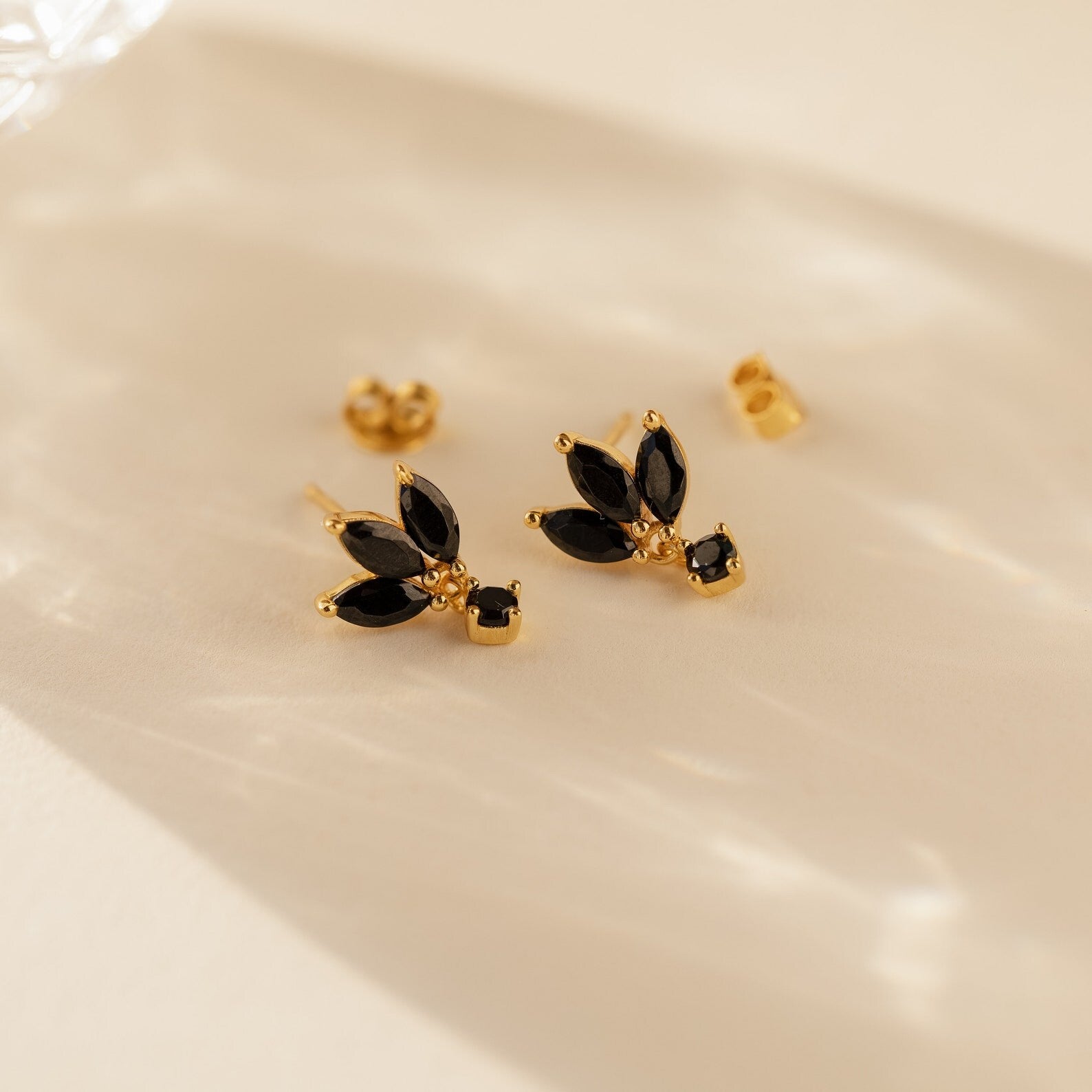 Emily Drop Earrings feature gold studs with black stones in a floral design, exuding vintage charm. These black petal earrings are beautifully displayed on a light surface.