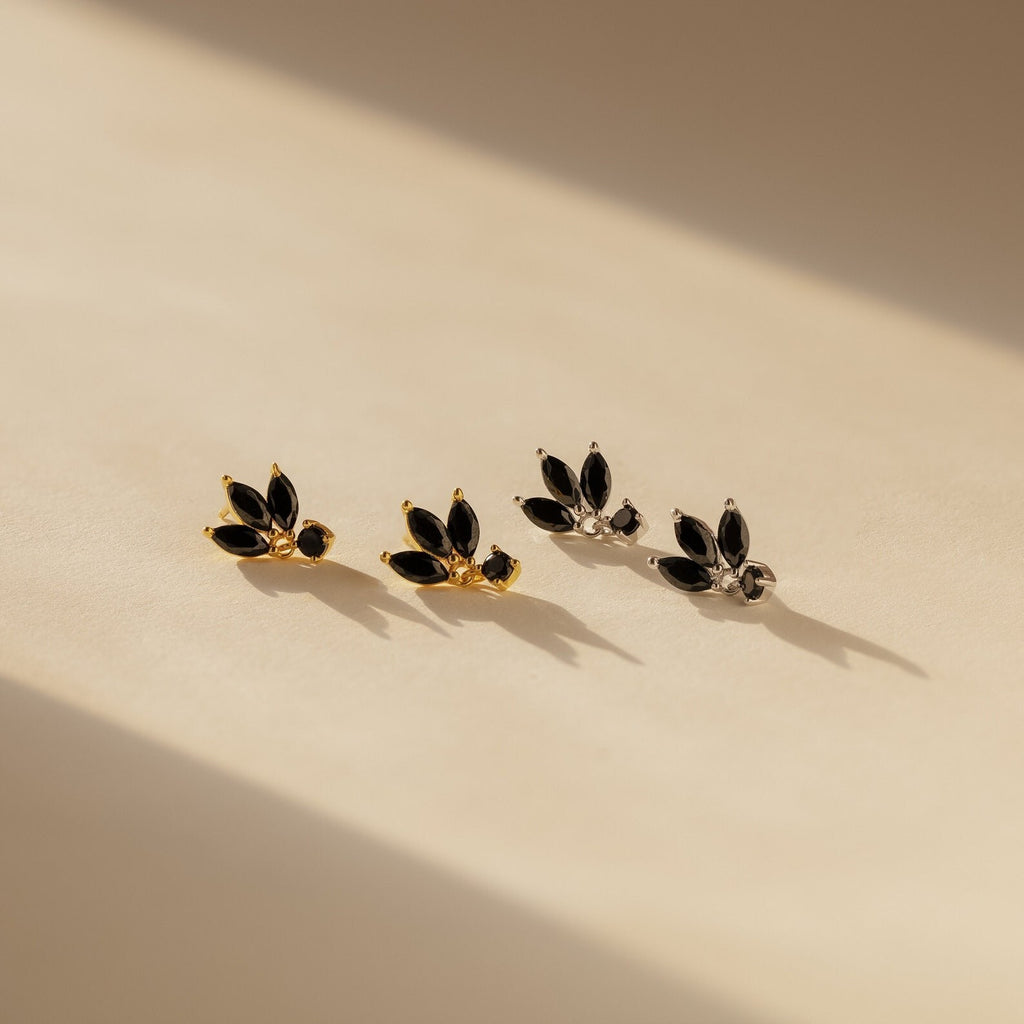 Emily Drop Earrings: two pairs—one gold, one silver—with black marquise stones evoking black petals, displayed on a beige surface.