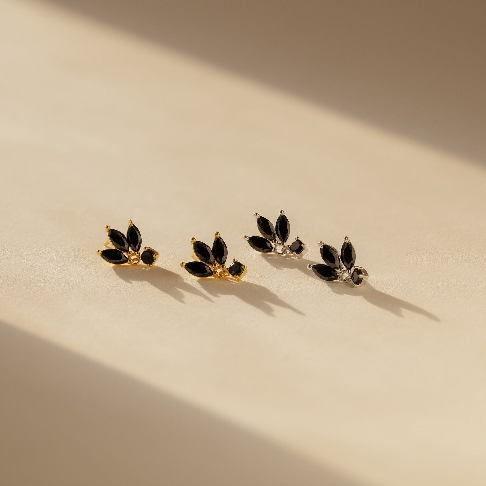 Emily Drop Earrings: two pairs—one gold, one silver—with black marquise stones evoking black petals, displayed on a beige surface.