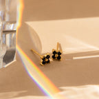 The Black Flower Studs, vintage earrings with gold and black stone accents, rest on a beige surface in sunlight, with a rainbow prism effect and a transparent crystal partially visible on the left.