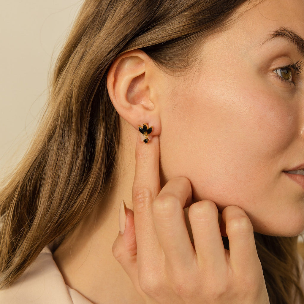 A woman touches her ear, showcasing the Emily Drop Earrings—small black and gold earrings with a flower-shaped, black petal design—in a close-up view.