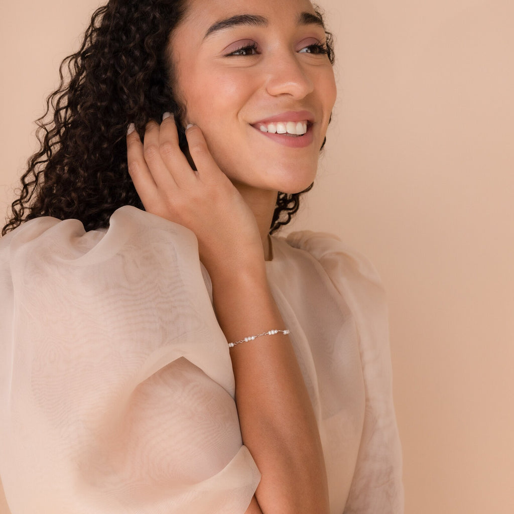 Smiling woman with curly hair wearing a sheer puff-sleeve blouse and the Hali Pearl Station Bracelet, perfect as bridal jewelry or a thoughtful bridesmaid gift.