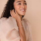 Smiling woman with curly hair wearing a sheer puff-sleeve blouse and the Hali Pearl Station Bracelet, perfect as bridal jewelry or a thoughtful bridesmaid gift.