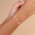 A hand displays the Hali Pearl Station Bracelet—delicate gold with small white pearls—against a beige background, making it an elegant choice for bridesmaid gifts or timeless bridal jewelry.