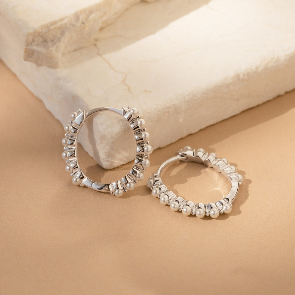The Malik Pearl Hoops, elegant silver earrings featuring small decorative beads, are ideal as minimalist accessories or chic wedding jewelry, displayed on a beige stone surface.