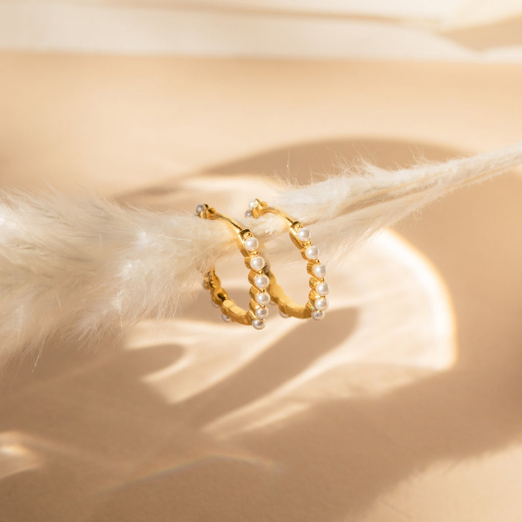 The Malik Pearl Hoops feature gold hoops adorned with small pearls, displayed on a soft beige surface under gentle lighting—an elegant choice for minimalist earrings or wedding jewelry.