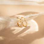 The Malik Pearl Hoops feature gold hoops adorned with small pearls, displayed on a soft beige surface under gentle lighting—an elegant choice for minimalist earrings or wedding jewelry.