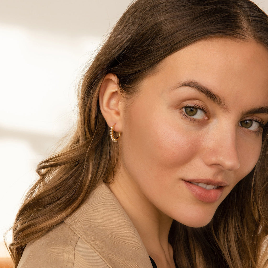 Woman with wavy brown hair smiles softly at the camera, wearing a beige jacket and Malik Pearl Hoops.