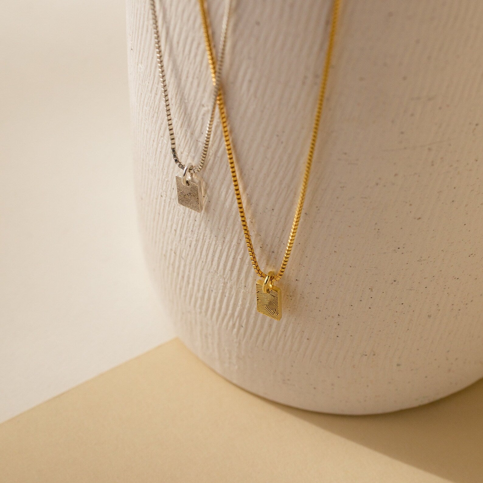 Two Mini Fingerprint Tag Necklaces, one silver and one gold, hang on a textured white ceramic vase—a meaningful personalized necklace and thoughtful Mother’s Day gift.