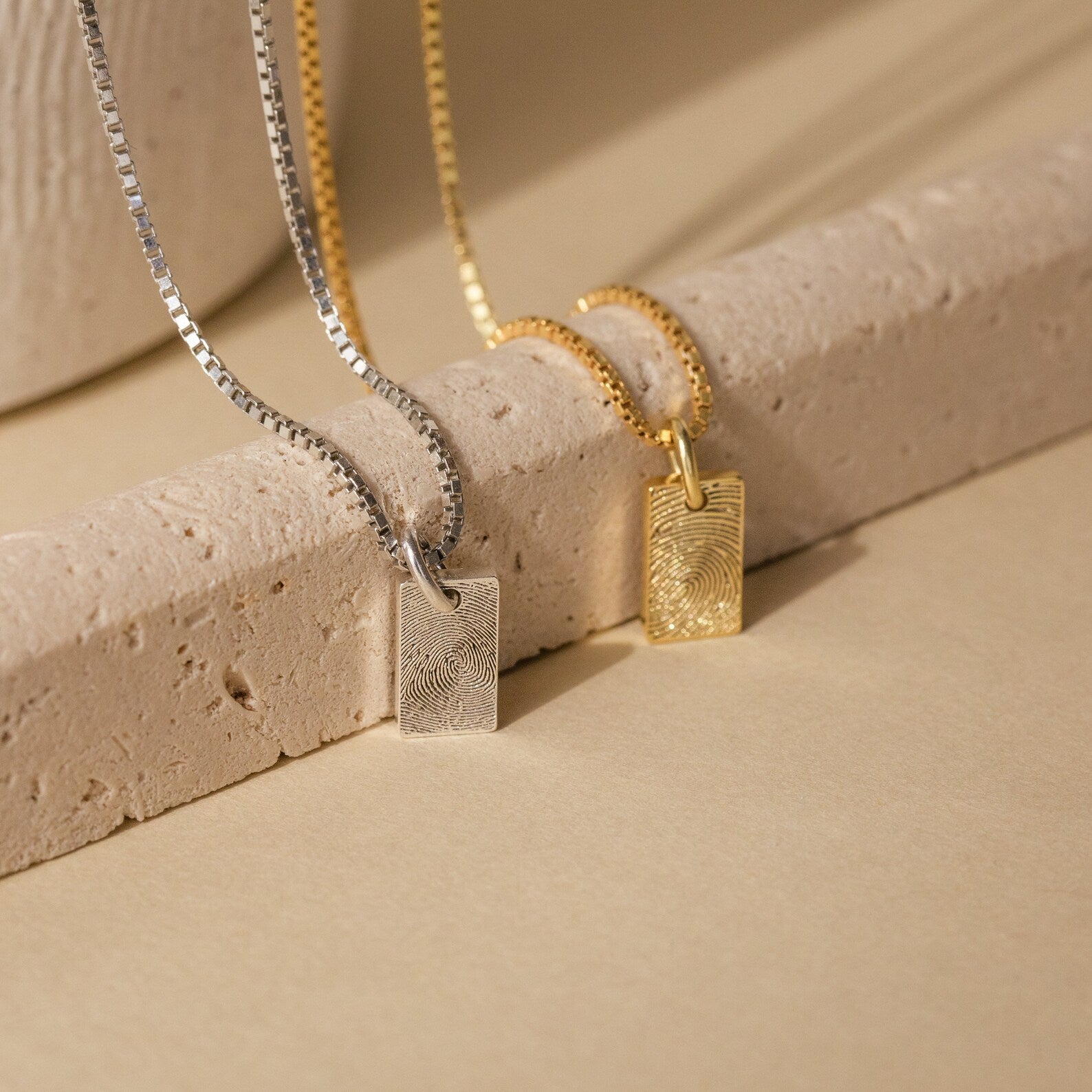 Two Mini Fingerprint Tag Necklaces with rectangular pendants, one silver and one gold, displayed on a beige stone—the perfect personalized Mother's Day gift.