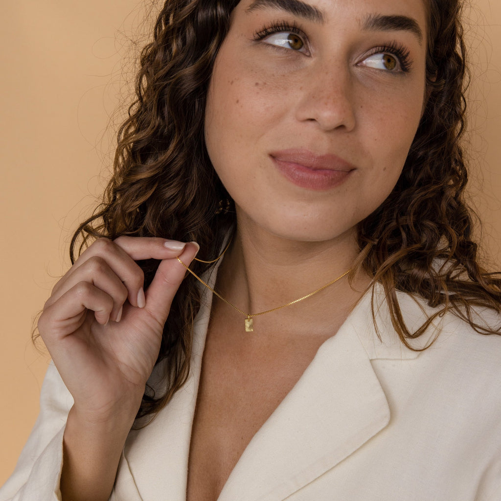 Woman with curly hair in a cream blazer smiles while holding the Mini Fingerprint Tag Necklace against a beige background—a thoughtful and unique Mother’s Day gift.