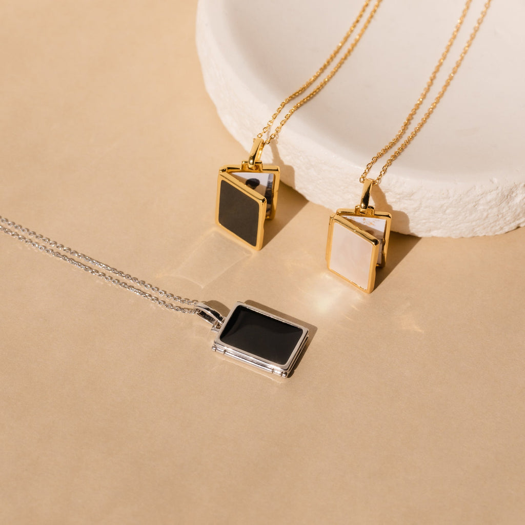 Three rectangular pendant necklaces, featuring the Onyx Locket Necklace and a sleek silver design, displayed on a beige surface and white dish—perfect as a personalized gift.
