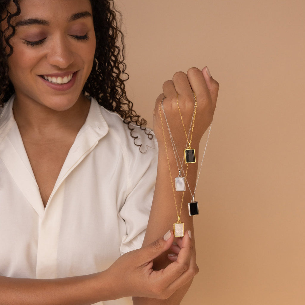A smiling woman in a cream blouse holds three gold and silver pendant necklaces, featuring the Onyx Locket Necklace, against a beige background.