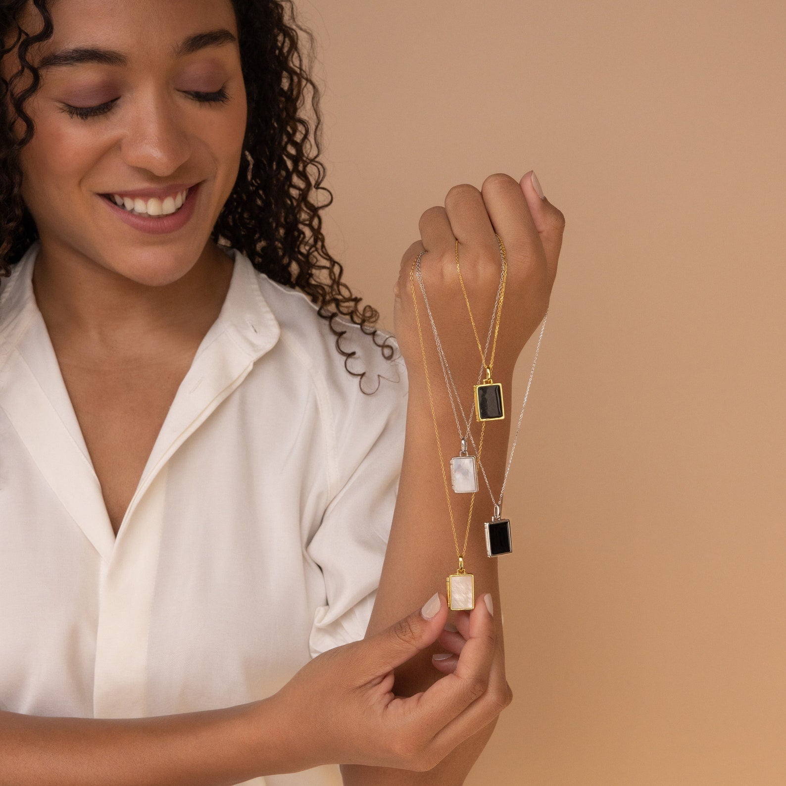 A smiling woman in a cream blouse holds three gold and silver pendant necklaces, featuring the Onyx Locket Necklace, against a beige background.