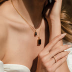 Woman wearing layered gold necklaces, including the Onyx Locket Necklace with a black enamel pendant and a thin gold ring, manicured nails, and a white off-shoulder top—an elegant look ideal for a personalized gift.