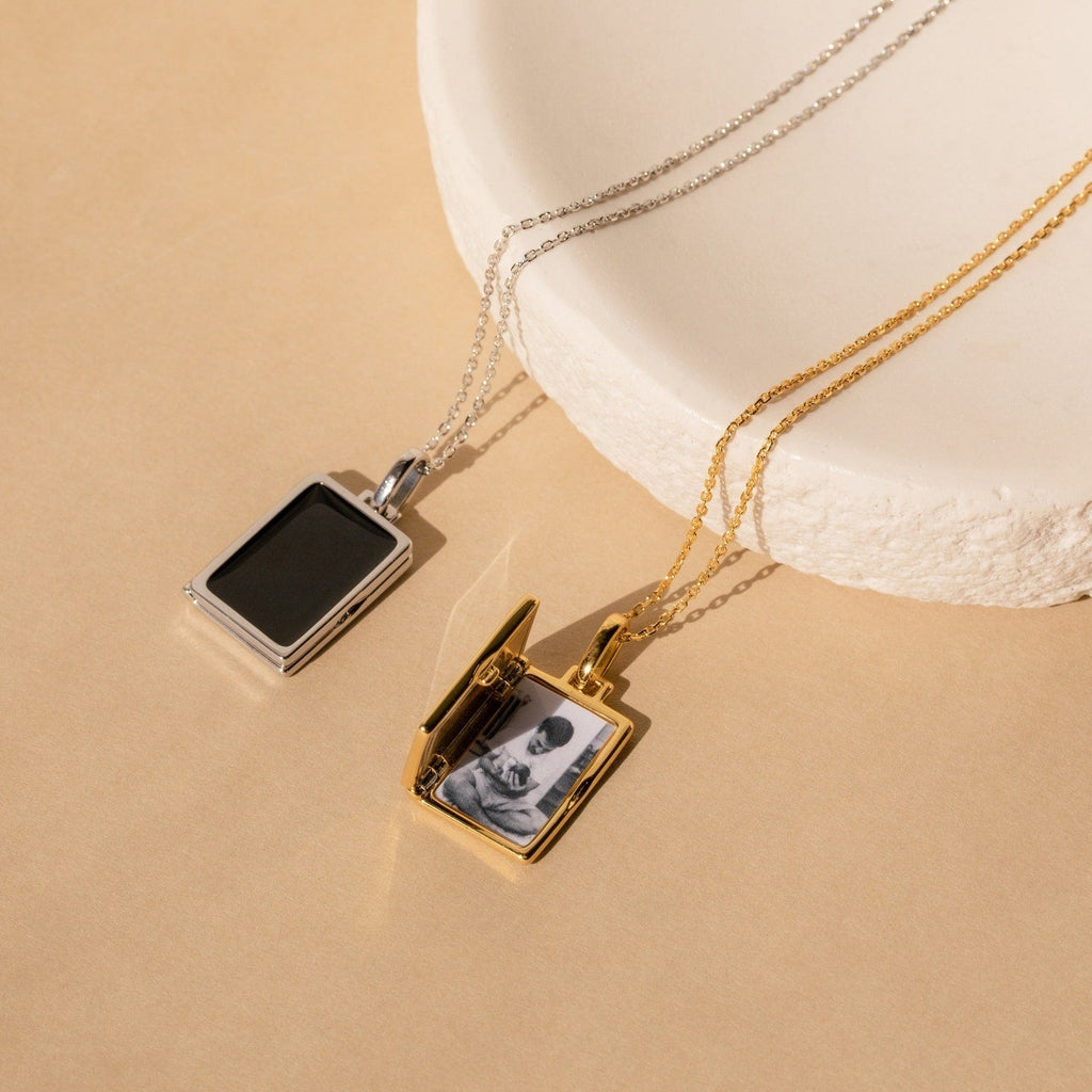 The Onyx Locket Necklace features two rectangular lockets—one in silver, one in gold—displayed on a beige surface. The gold locket charm is open, making it a thoughtful, personalized gift.