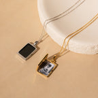 The Onyx Locket Necklace features two rectangular lockets—one in silver, one in gold—displayed on a beige surface. The gold locket charm is open, making it a thoughtful, personalized gift.