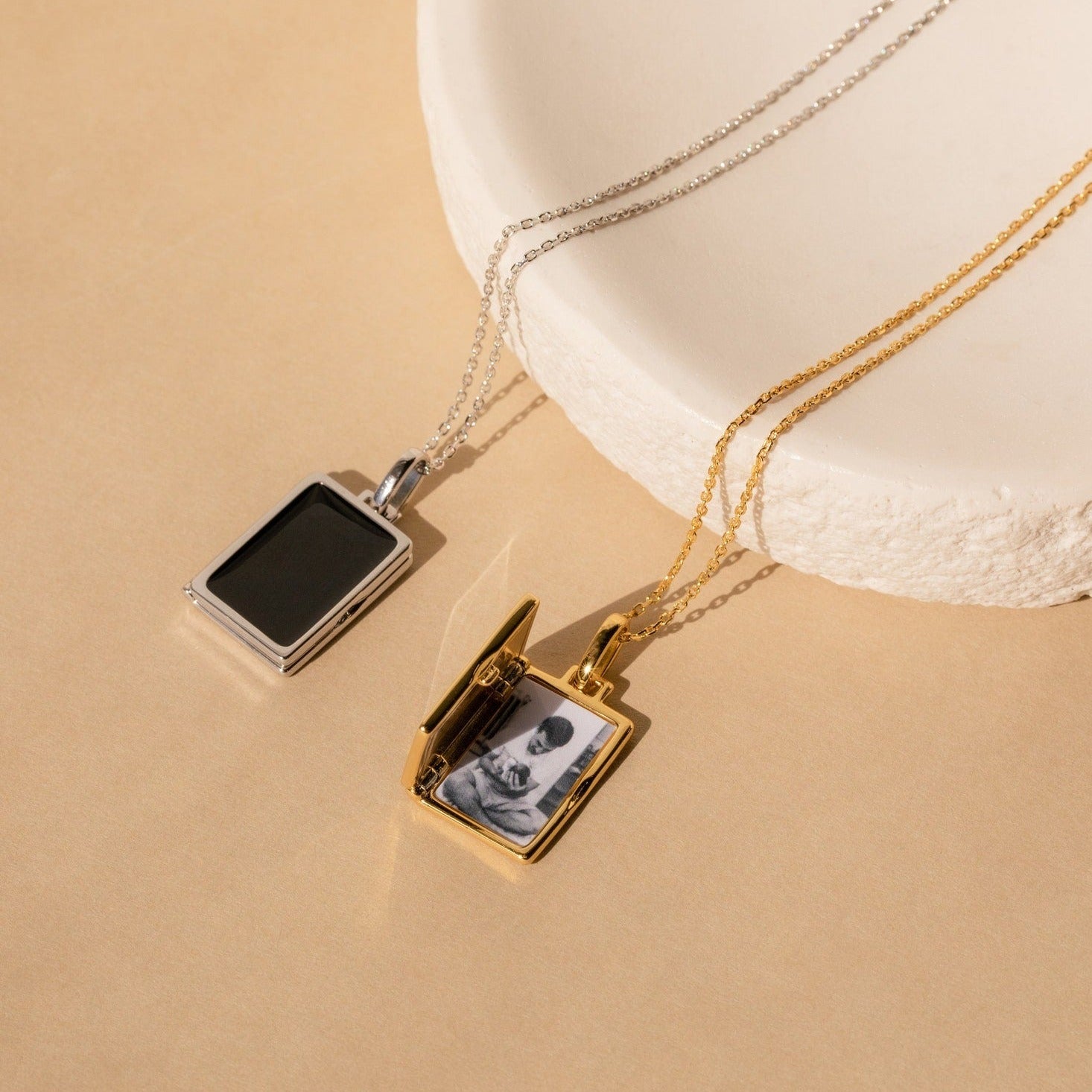 The Onyx Locket Necklace features two rectangular lockets—one in silver, one in gold—displayed on a beige surface. The gold locket charm is open, making it a thoughtful, personalized gift.