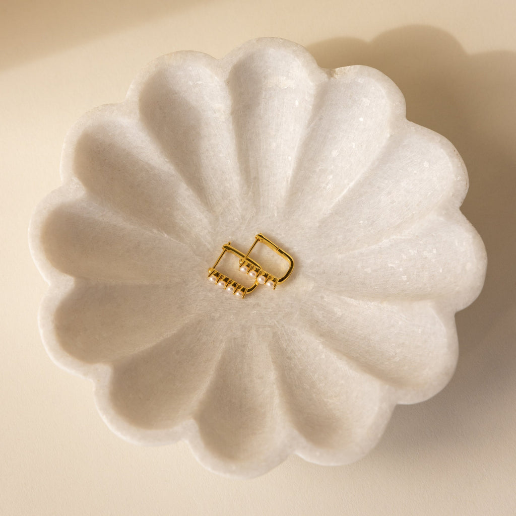 The Rayne Pearl Link Earrings—gold hoops with freshwater pearls—are displayed in a white scalloped dish on a beige surface, perfect for minimalist jewelry lovers.