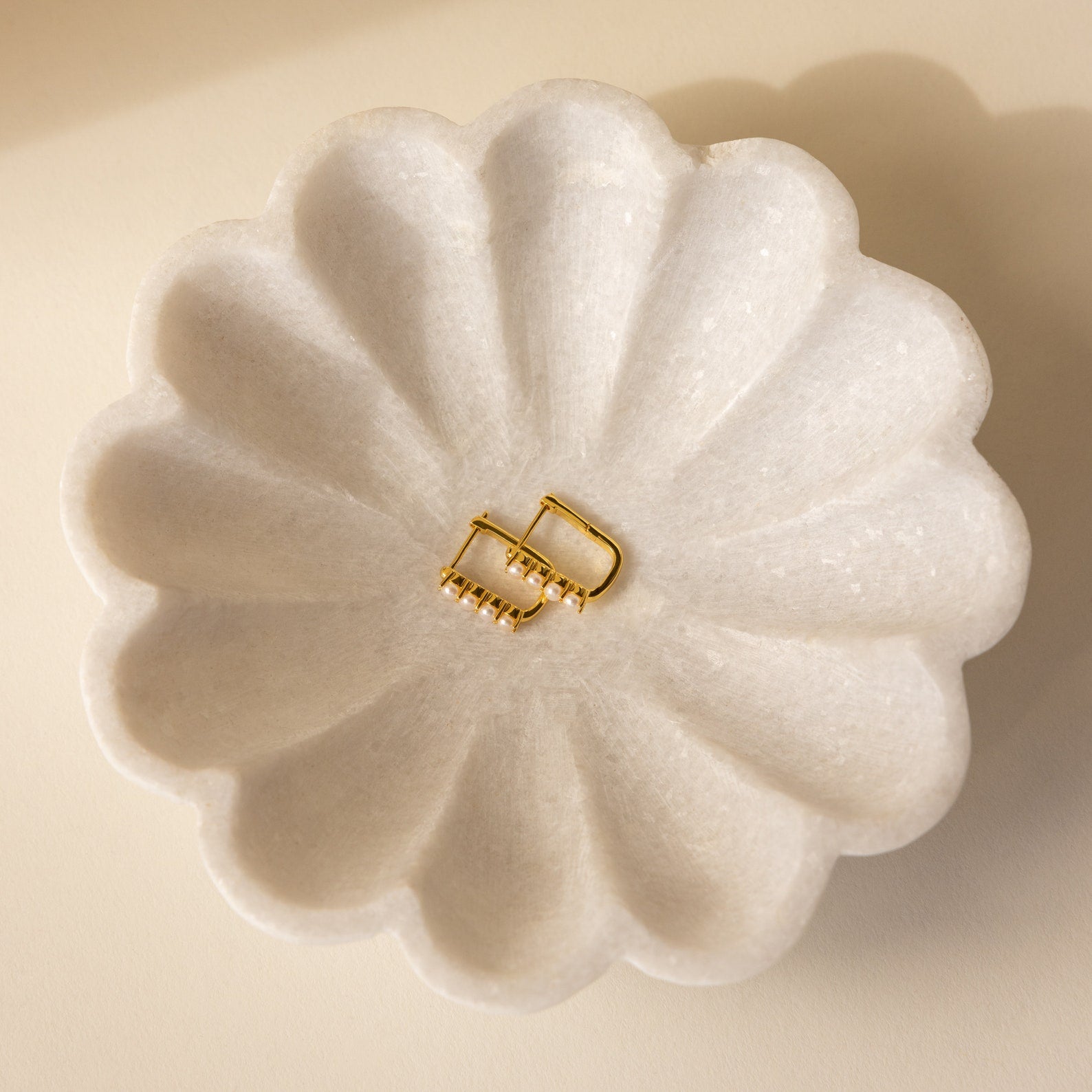 The Rayne Pearl Link Earrings—gold hoops with freshwater pearls—are displayed in a white scalloped dish on a beige surface, perfect for minimalist jewelry lovers.