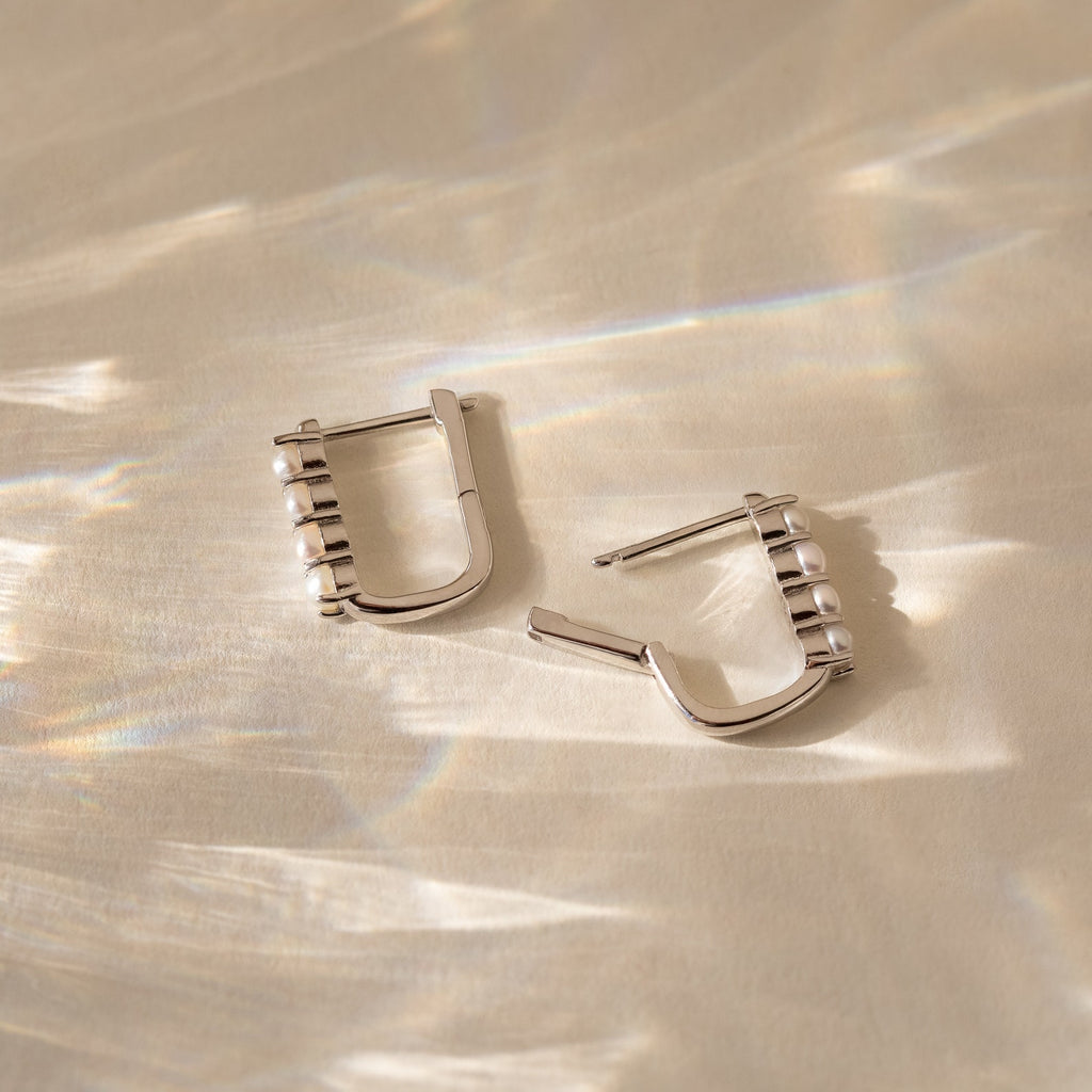 The Rayne Pearl Link Earrings feature square silver links adorned with small freshwater pearls, displayed on a beige surface with reflected light—ideal for fans of minimalist jewelry.