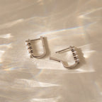 The Rayne Pearl Link Earrings feature square silver links adorned with small freshwater pearls, displayed on a beige surface with reflected light—ideal for fans of minimalist jewelry.