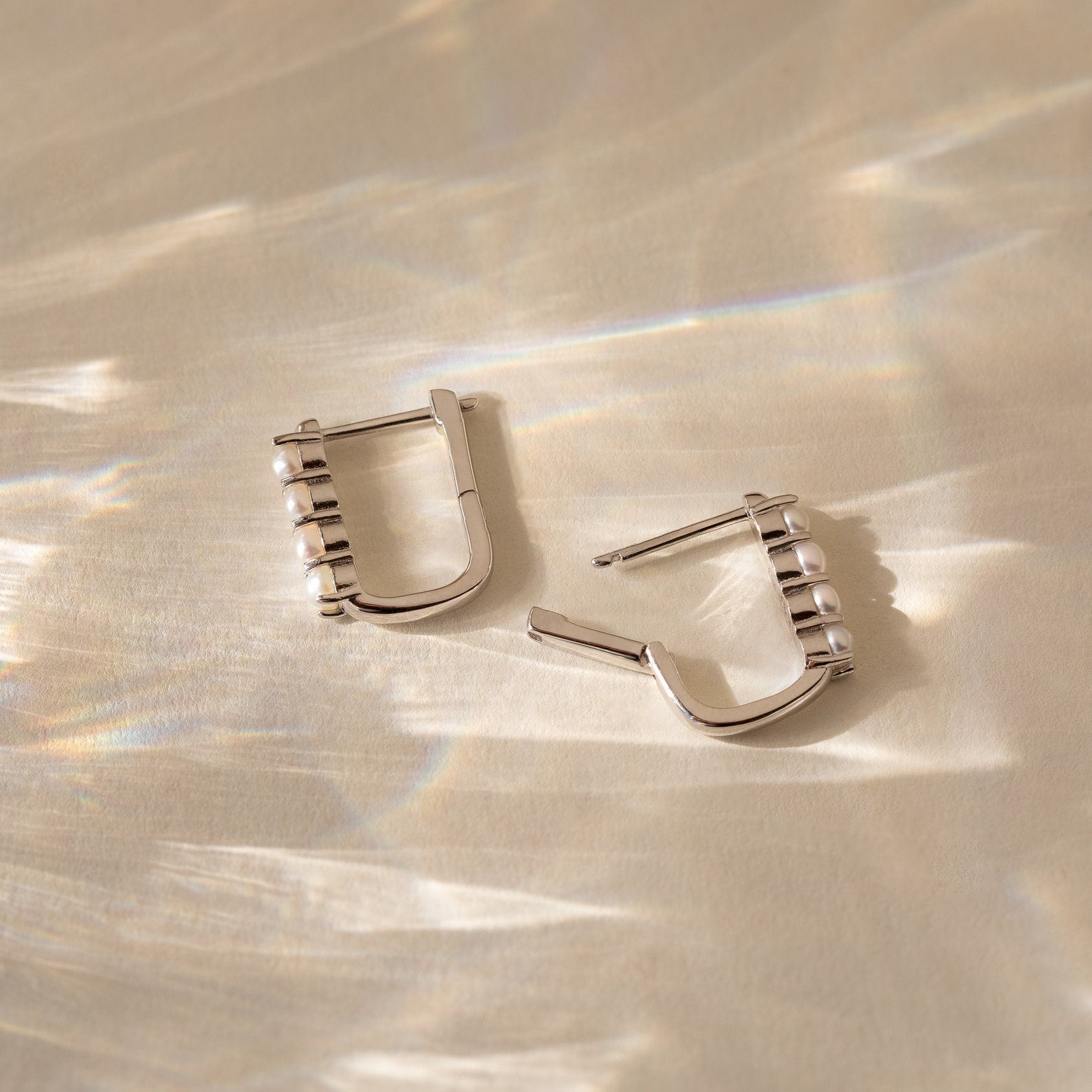 The Rayne Pearl Link Earrings feature square silver links adorned with small freshwater pearls, displayed on a beige surface with reflected light—ideal for fans of minimalist jewelry.