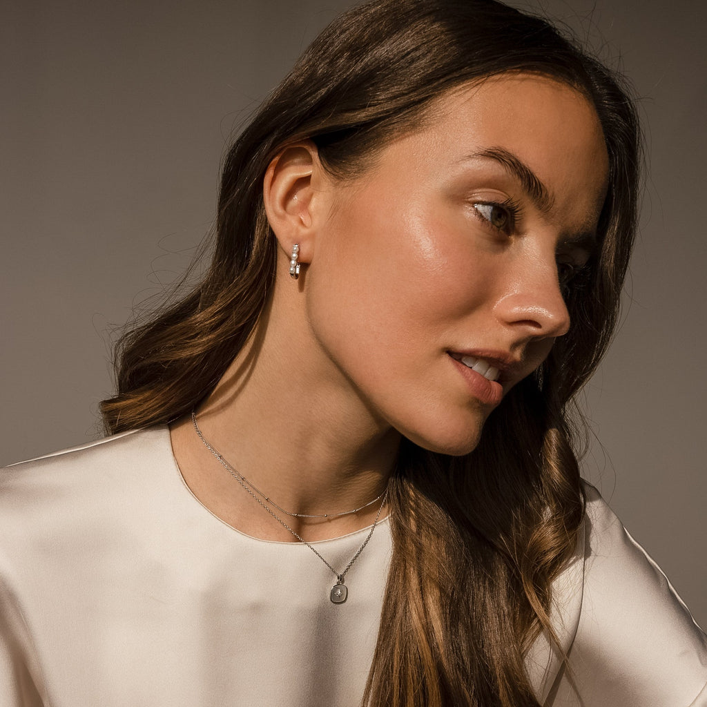 A woman with long brown hair wears minimalist jewelry—Rayne Pearl Link Earrings and a silver necklace—paired with a cream-colored top.