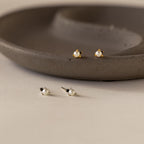 Two pairs of pearl stud earrings—one with gold, one with silver accents—are displayed by a ceramic dish. Effortlessly pair them with our Raindrop Pearl Onyx Studs for timeless elegance.
