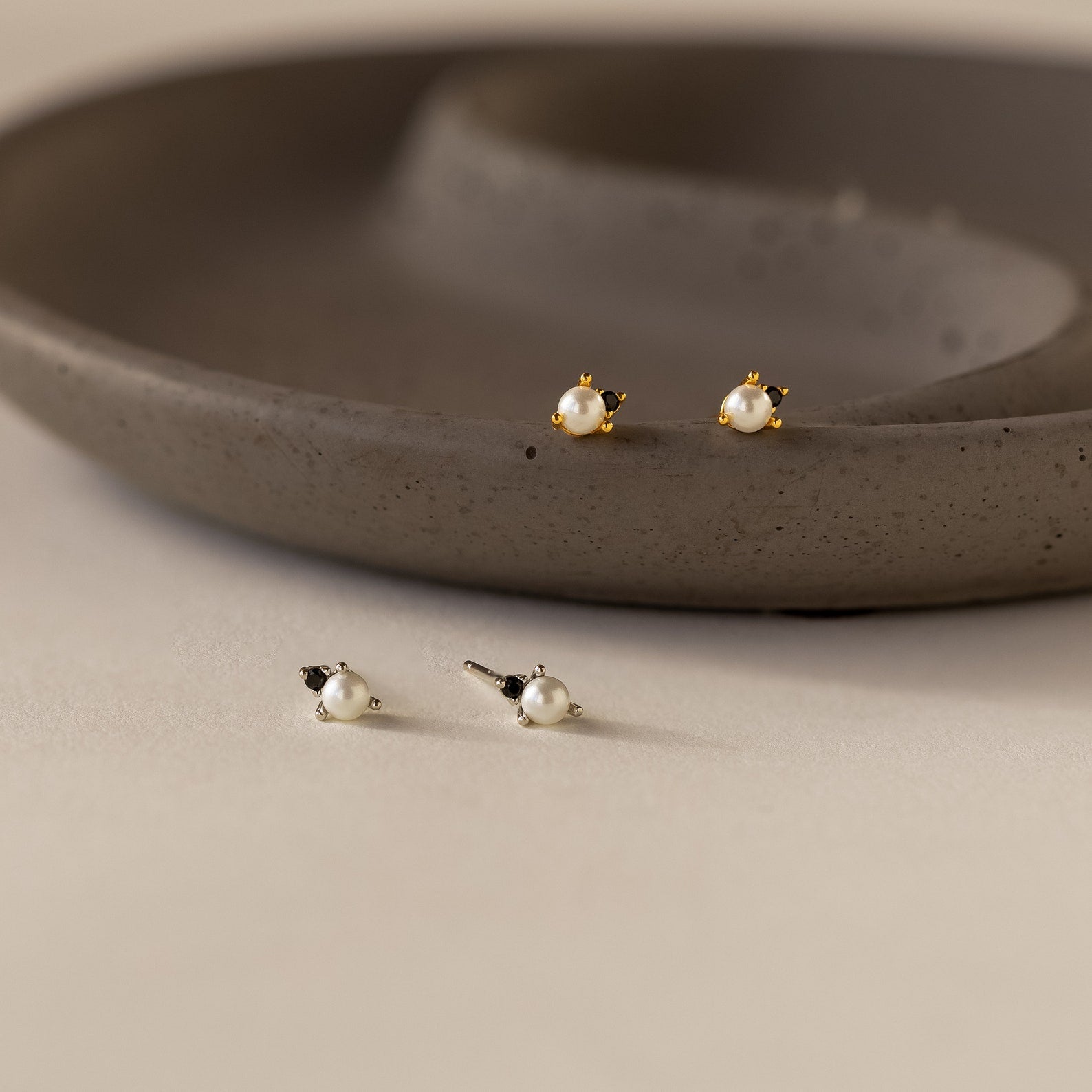 Two pairs of pearl stud earrings—one with gold, one with silver accents—are displayed by a ceramic dish. Effortlessly pair them with our Raindrop Pearl Onyx Studs for timeless elegance.
