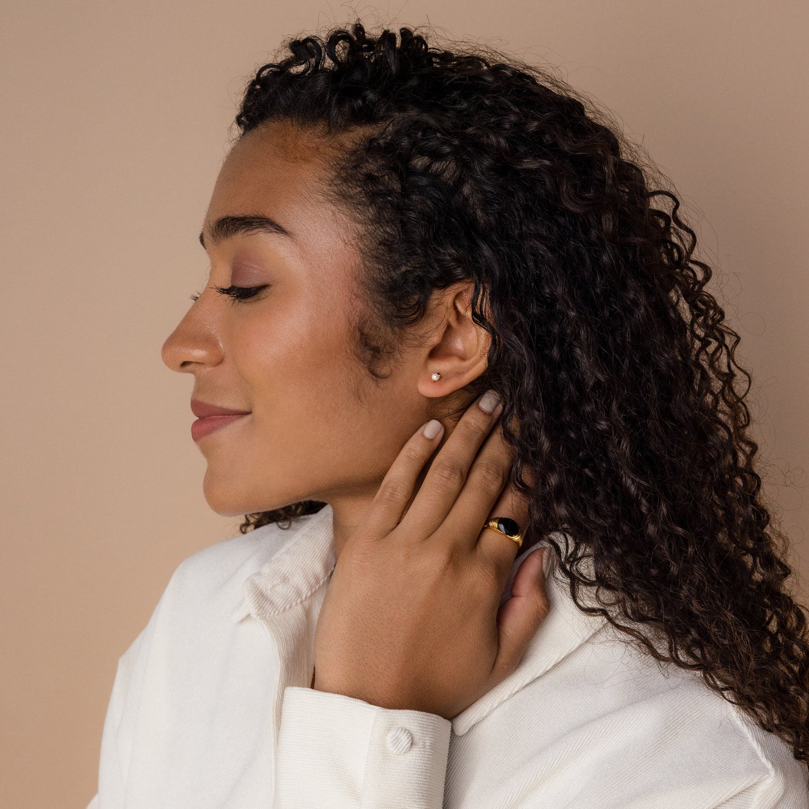 Woman with curly hair in profile, eyes closed, touching her neck, wearing a white shirt, gold ring, and elegant Raindrop Pearl Onyx Studs.