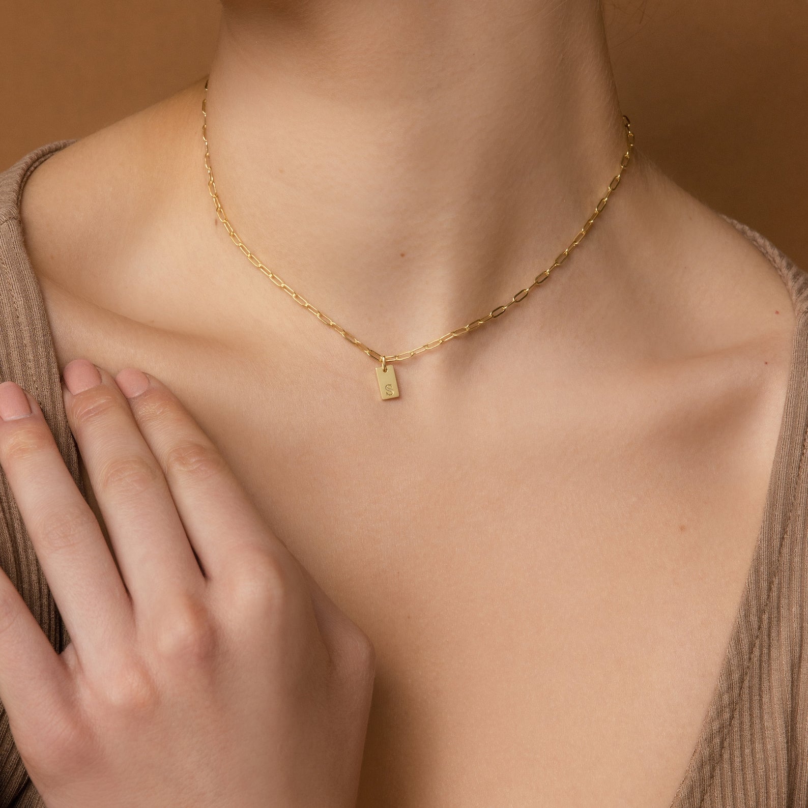 Woman wearing the Mini Initial Paperclip Necklace, featuring a gold chain and small rectangular initial pendant, gently touches her collarbone—a delicate, personalized piece perfect for everyday elegance.