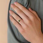 A hand adorned with the Duo Vintage Birthstone Ring—featuring a gold finish, a green stone, and a small white gem—rests on green fabric, making for an elegant Mother’s Day gift.