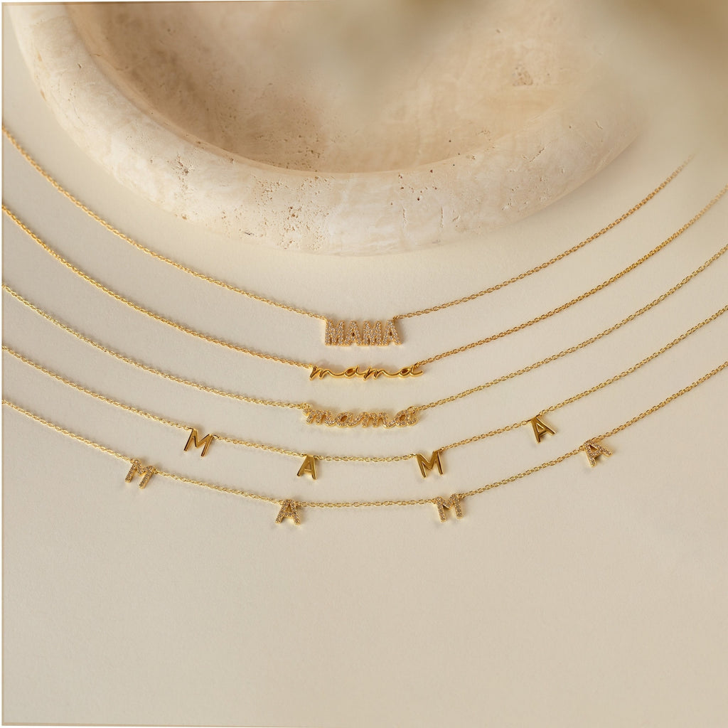Five gold Mama Necklaces, each featuring a distinct letter style, are displayed on a beige surface near a round stone dish—an elegant and thoughtful gift for Mom, crafted to celebrate every moment.
