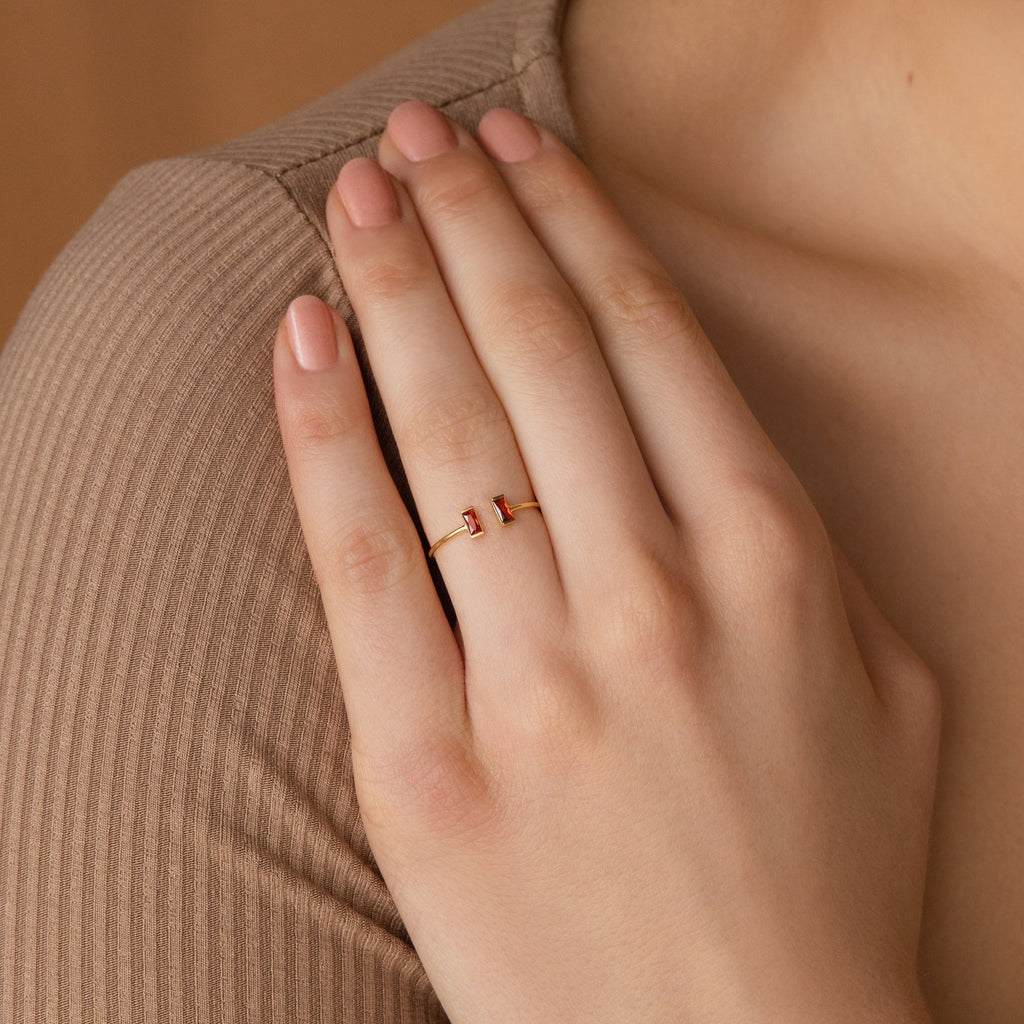 A hand with nude nails wears the Duo Vertical Baguette Ring—a delicate gold ring with two small red stones on the ring finger, making it a perfect personalized gift.