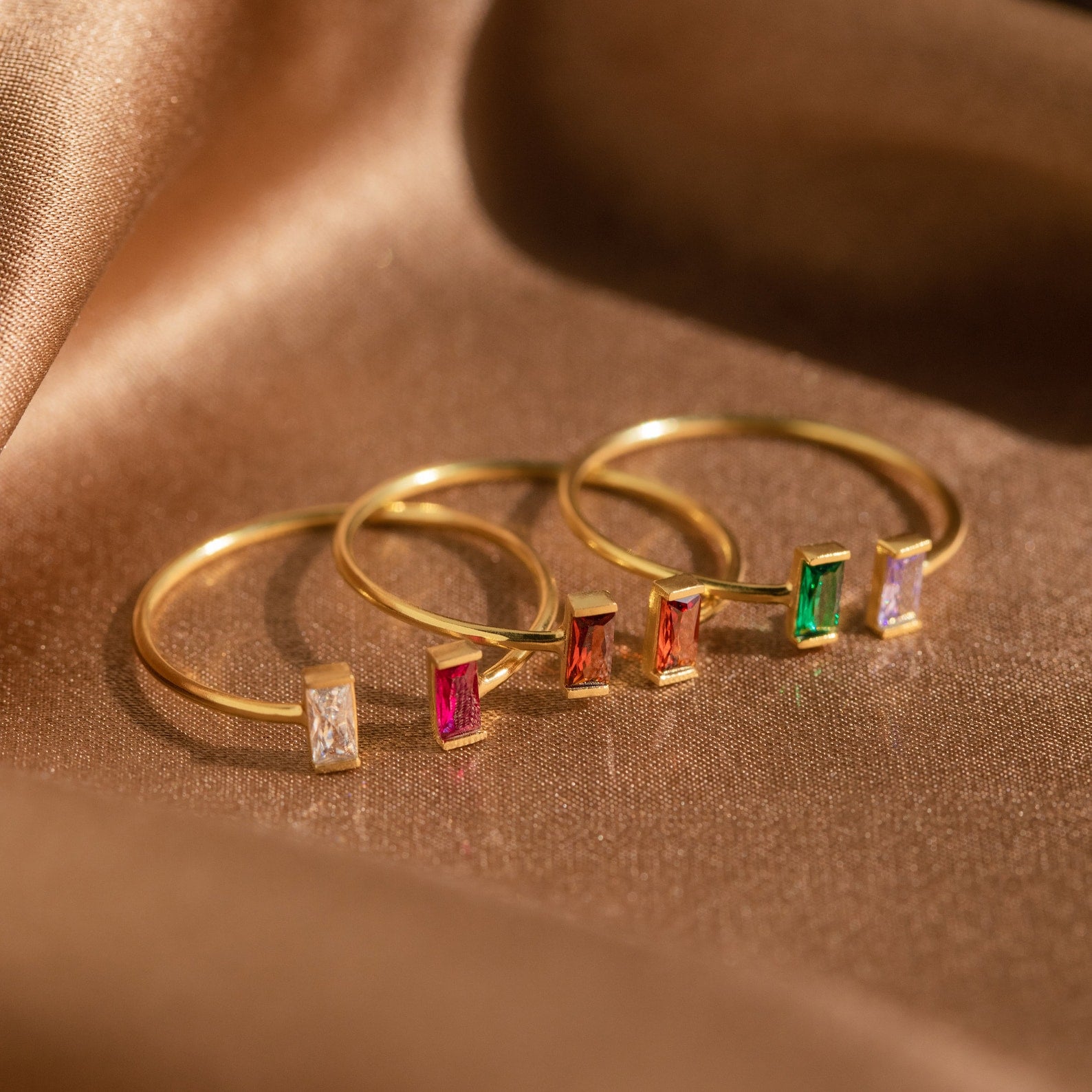 Four Duo Vertical Baguette Rings with rectangular gemstones in white, pink, red, green, and lavender rest on brown satin fabric—a stunning personalized gift or custom birthstone ring for someone special.