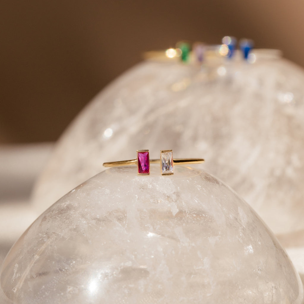 The Duo Vertical Baguette Ring features two rectangular stones, one pink and one clear, set on gold and displayed atop a crystal dome—a stunning custom birthstone ring, perfect as a personalized gift.