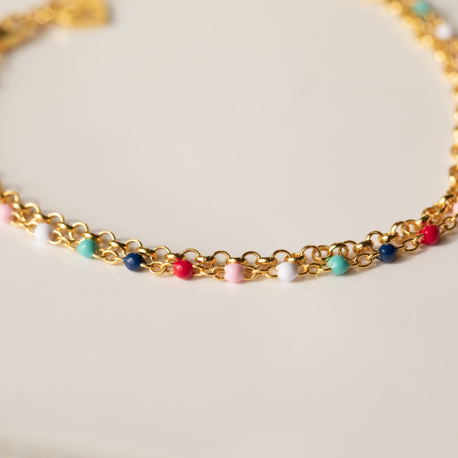 Rainbow Station Bracelet features a gold double chain decorated with small, colorful beads in pink, blue, white, red, and turquoise against a light background.