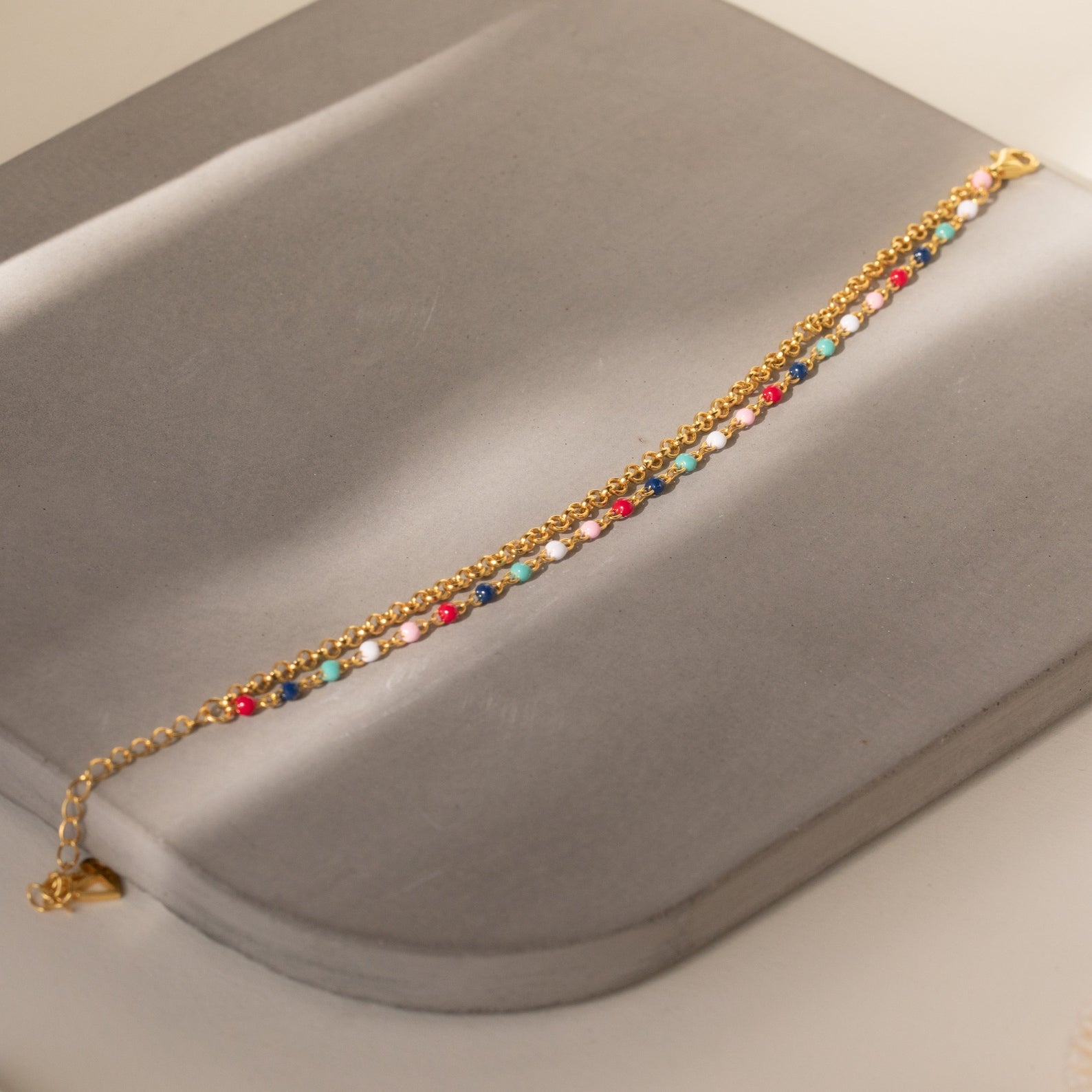 The Rainbow Station Bracelet is a handmade double chain bracelet with colorful beads, shown on a gray, curved-edged surface in soft lighting.