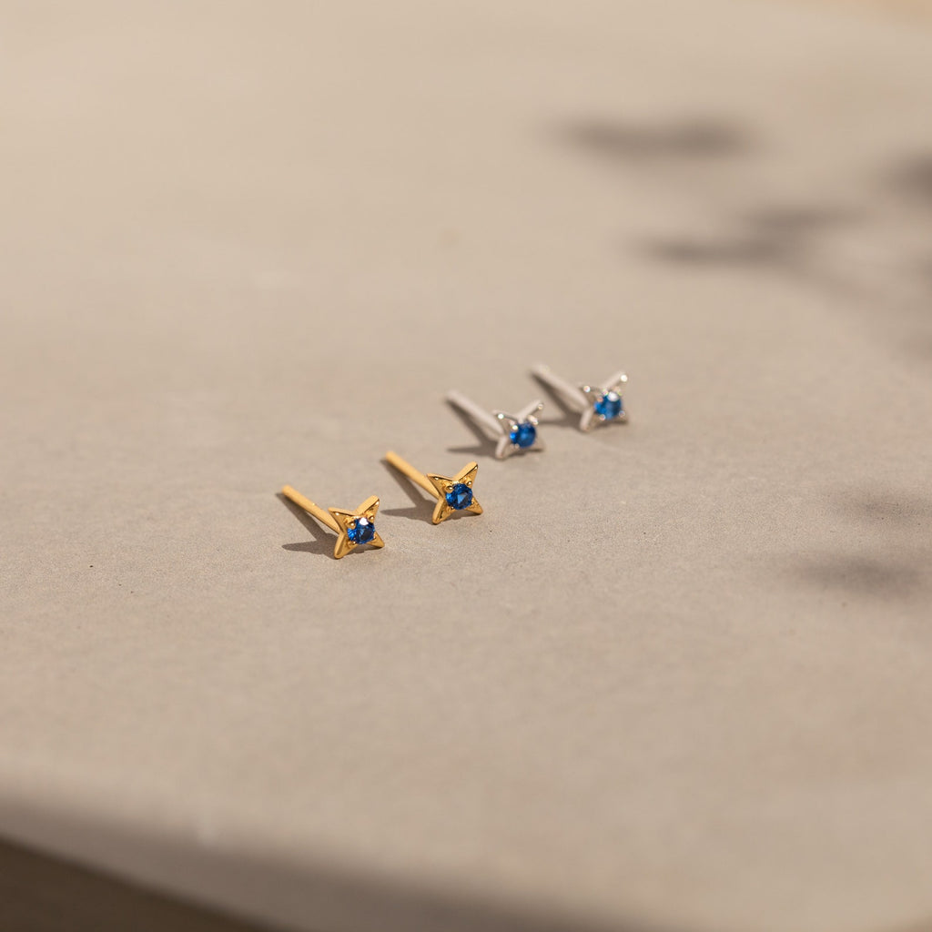 Two pairs of Sapphire Star Studs, one gold and one silver with small sapphire gemstones—ideal as Sapphire Star Stud Earrings or for cartilage piercings—are displayed elegantly on a beige surface.