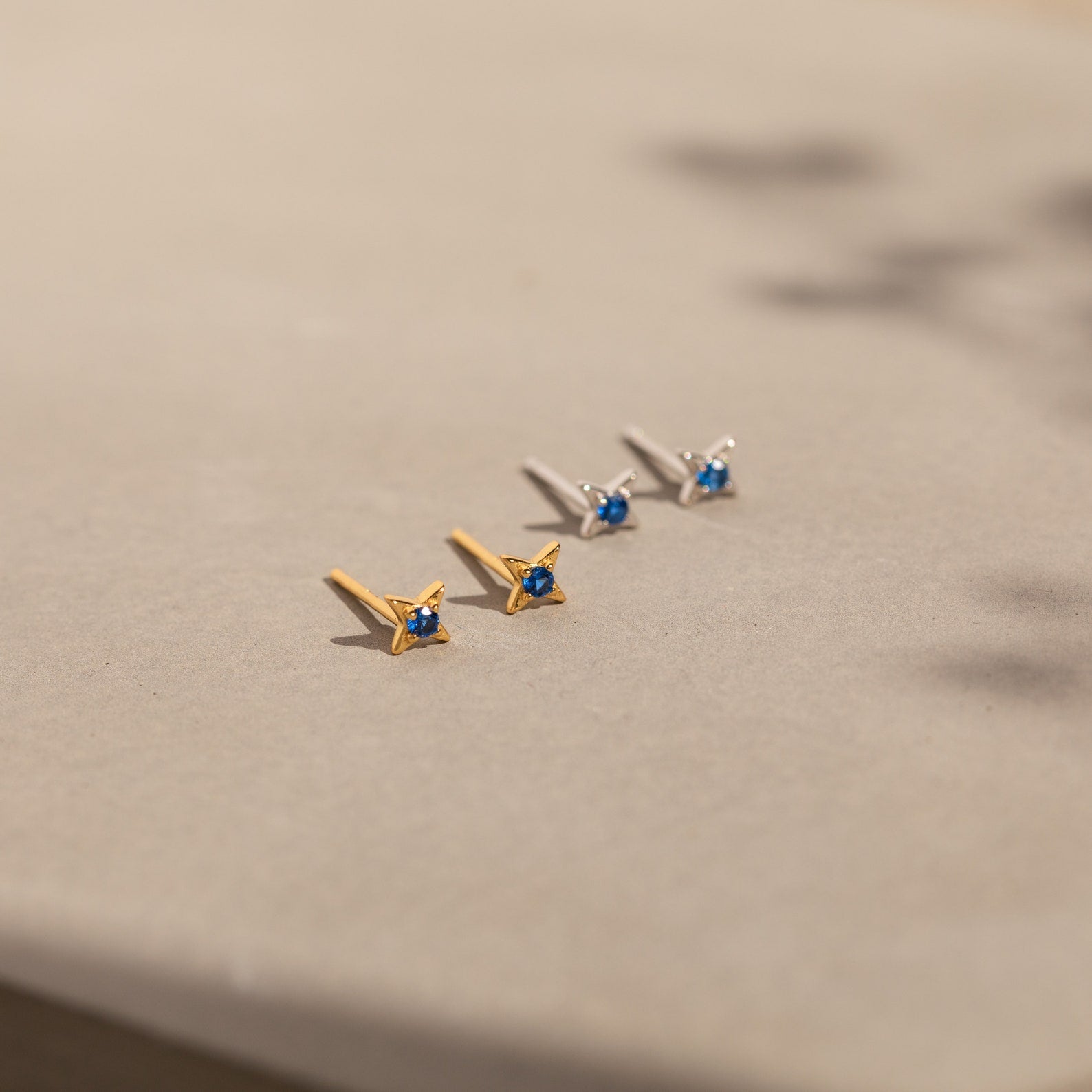 Two pairs of Sapphire Star Studs, one gold and one silver with small sapphire gemstones—ideal as Sapphire Star Stud Earrings or for cartilage piercings—are displayed elegantly on a beige surface.
