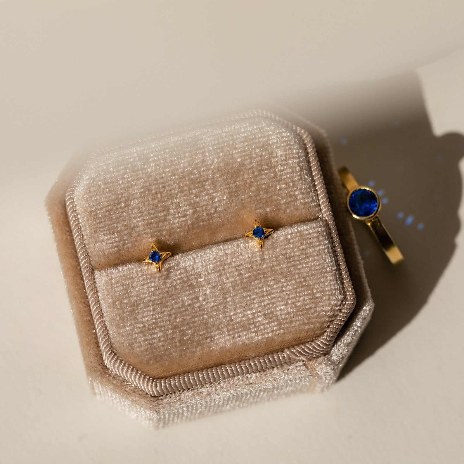 Sapphire Star Studs and a gold ring with blue stones are displayed on a beige velvet jewelry box in soft lighting.