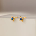 Sapphire Star Studs: Gold star-shaped earrings with sparkling blue gems, perfect for cartilage piercings or daily wear. Displayed on a white surface with a soft background, they add celestial charm to any look.