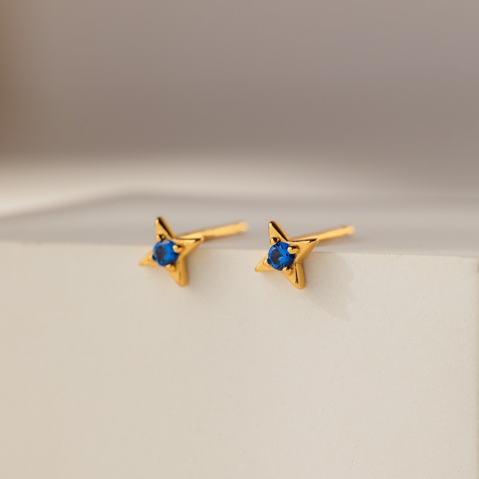 Sapphire Star Studs: Gold star-shaped earrings with sparkling blue gems, perfect for cartilage piercings or daily wear. Displayed on a white surface with a soft background, they add celestial charm to any look.
