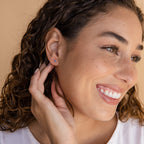 A smiling woman with curly hair wears Sapphire Star Studs and touches her ear with one hand.