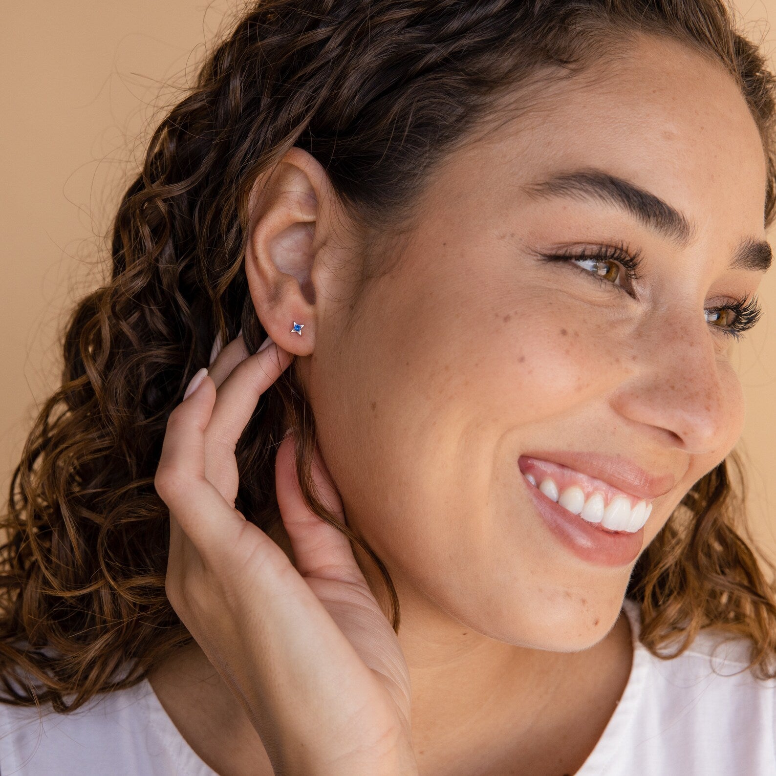 A smiling woman with curly hair wears Sapphire Star Studs and touches her ear with one hand.